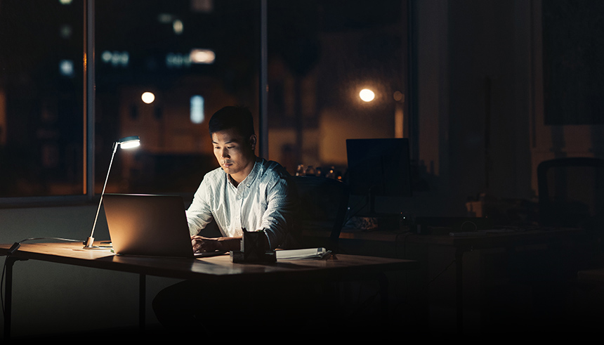 Young Asian businessman working on a laptop while sitting at his desk in a dark office at night with city lights in the background